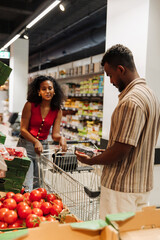 A man reads the package he is holding while a woman stands next to him and holds a cart
