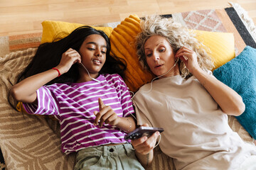 Mother showing phone to her daughter while they listen to earphones and lie on pillows and blankets