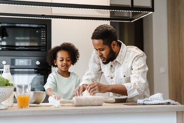 Father talking and picking up dough with his smiling daughter while they stand at the table