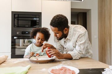 Daughter holds a spoonful of sauce over dough in a baking dish while father stands next to her at the table holding a jar