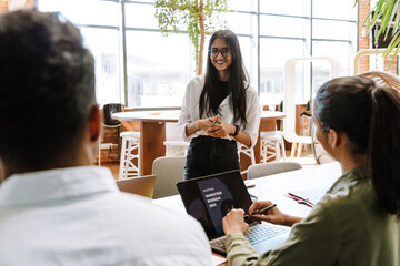 A female worker stands and smiles while listening to a female and male worker sitting across from her at a table