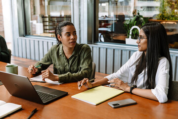 Fototapeta premium Two female workers talking and sitting at a table while holding a pencil and pen