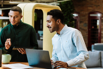 Two male workers are smiling and looking ahead while they are at a table and one of them is holding a computer tablet