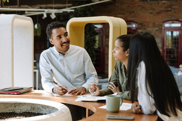 A male worker smiles and talks to two female workers while they sit at a table
