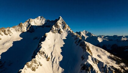 Dramatic aerial view of majestic snow-covered alpine mountain range with deep shadows and sharp ridges against a clear blue sky