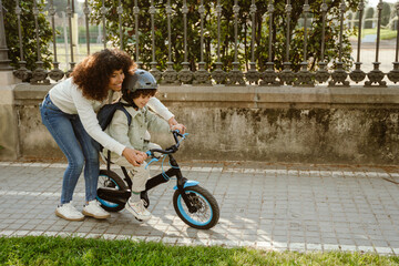 A mother walks bent over and puts her hands on the arms of her son who is riding a bicycle in front of her while they laugh