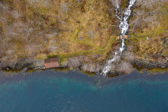 Top-down aerial view of a rugged lakeshore where a narrow waterfall meets clear blue water, a small wooden bridge and footpath winding through dry grass, bare trees, and mossy terrain. - Powered by Adobe