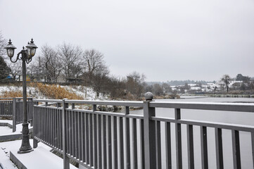 Winter riverside landscape with metal railing and vintage street lamps covered in snow. Snowfall over a calm river, bare trees, and rural houses in the background.