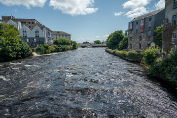 View of the river Corrib flowing through Galway, Ireland, with adjacent buildings and vibrant greenery under a partly cloudy sky.