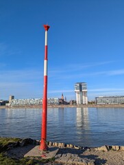 Bright red pole guides boats at shoreline, Red navigational post by water with city skyline reflection, Vivid red marker along riverbank beneath clear blue daylight sky