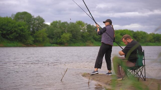 Romantic couple standing on the river bank and fishing with spinning rods during a cloudy day, enjoying their outdoor leisure activity and spending quality time together in nature