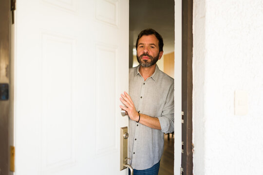 Latin man opening front door, cautiously looking out, welcoming visitors or homeowners arriving home