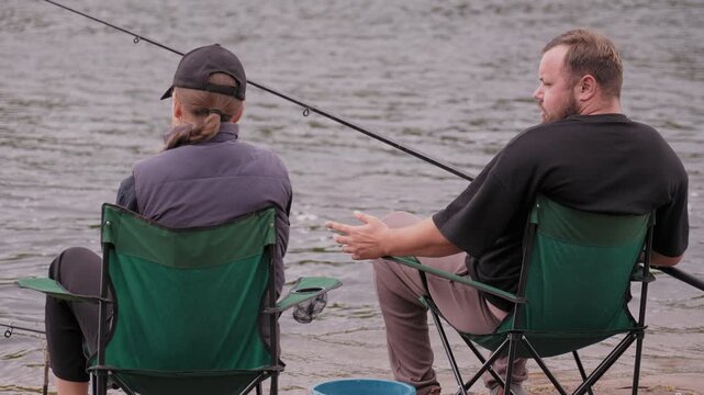 Middle-aged bearded man patiently teaching a young woman how to use a spinning reel on a fishing rod by the river, helping her untangle the fishing line for their joint hobby
