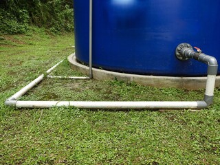 A large blue water storage tank sits on a concrete base with white PVC pipes running along the ground on a bright green, grassy lawn in a tropical setting.