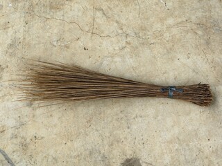 Traditional Indonesian or Southeast Asian broom Sapu Lidi made from bundled coconut palm leaf ribs, resting on a textured, cracked concrete floor. Rustic and eco-friendly cleaning tool.