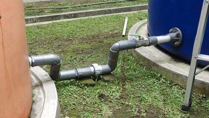 A gray PVC pipe connection, including a green ball valve, linking two large outdoor plastic water storage tanks orange and blue on a grassy area with concrete curbs.