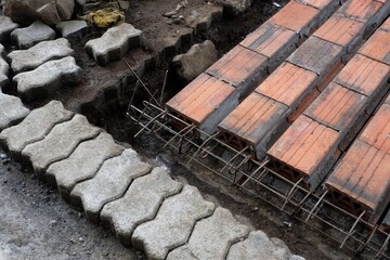 High-angle view of a construction base layer showing gray interlocking concrete pavers next to a trench with rebar framework and orange hollow bricks ready for a foundation pour.