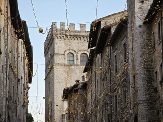 A narrow medieval street in the Italian town of Gubbio is decorated with warm Christmas lights, leading the eye towards the historic stone Palazzo dei Consoli