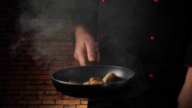 A chef tosses a pan of food as steam rises in the kitchen. The setting is dark, the wall is brick, and the cook is focused on the cooking process