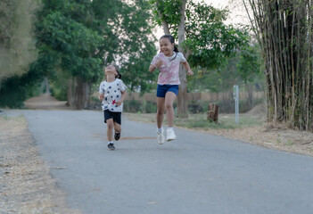 Obraz premium Two happy Asian children running and playing joyfully on an empty country road lined with bamboo. Focus on active childhood, freedom, summer fun, and energetic movement outdoors.