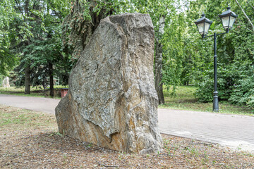 Large granite boulder stands on gravel in a park beside a street lamp. Green trees and shadows create a calm atmosphere. © Viacheslav