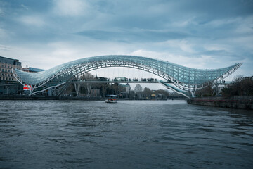 The Bridge of Peace in Tbilisi, Georgia, features a modern bow-shaped glass and steel canopy spanning over the Kura River under a cloudy sky © Contentino
