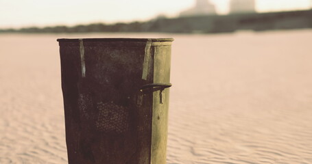 A rusty trash can stands alone on a sandy beach during sunset. The warm light highlights its weathered surface, contrasting with the peaceful surroundings, hinting at human impact on nature.