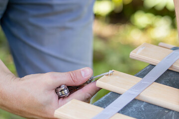 A close-up of a man's hand working with wooden planks and tools in a garden. He uses a multi-tool to remove staples from the slats.