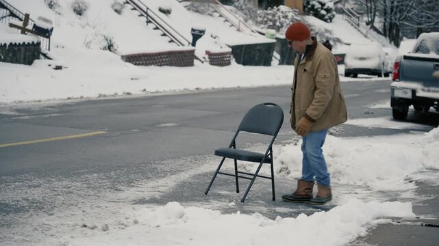 A wide shot view of a man placing a folding chair in a cleared street parking space, reflecting the Pittsburgh tradition of reserving spots after shoveling snow. 14979  