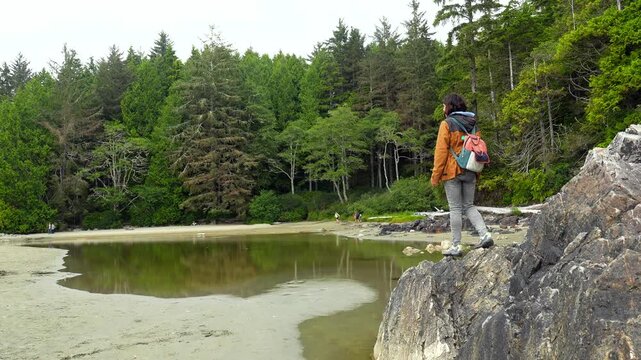 Female hiker with a backpack carefully balances on a rock at mackenzie beach in tofino, vancouver island, british columbia, canada, enjoying the serene view of the ocean and lush forest