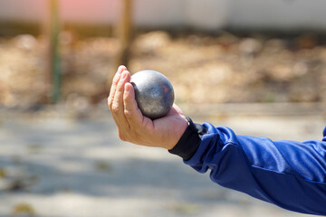 A person's hand, wearing a blue sleeve, is holding a silver metal p&eacute;tanque (bocce) boule, demonstrating the grip and preparation before throwing the ball in this outdoor sport.