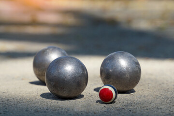 Three silver metal pétanque (bocce) boules surround a small, colored cochonnet (jack) ball on a gravel court, representing the scoring and challenge phase of the game.