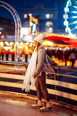 A young woman skates against the backdrop of a town square, decorated with bright holiday lights. A...