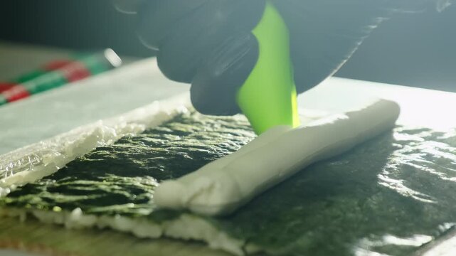 Chef hand in black gloves squeeze soft cream cheese onto nori with rice on mat closeup. Asian cuisine master prepares tasty sushi in kitchen