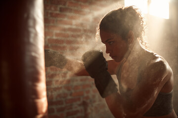 Hispanic female boxer striking a heavy bag in a sunlit warehouse gym. Powerful, sweaty, and authentic boxing fitness energy. Female athlete, woman training for power and muscles. Healthy lifestyle.