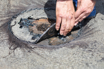 Worker lifts deteriorated roofing material during flat roof drainage system repair, exposing rusted metal and tar-coated layers. Closeup highlights inspection and maintenance of damaged roof component © Leilani
