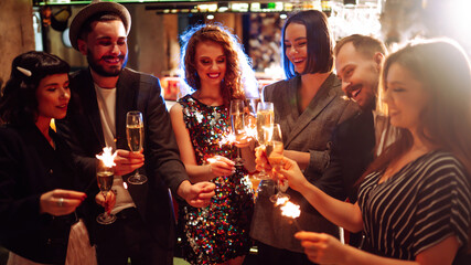 Young friends having fun with sparklers and glasses of champagne at corporate party. Handsome men and woman spend time together, celebrating New Year in cozy space. Сoncept is festive and fun.