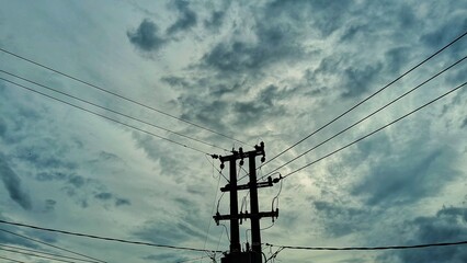 Fototapeta premium A low-angle, silhouette view of a power pole and electrical wires against a dramatically cloudy, dark sky, giving an ominous mood.