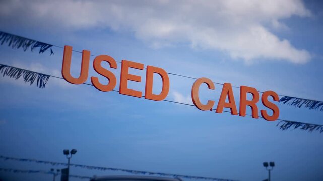 Used cars, a sign and streamers for a used car dealership in a suburban community against a blue sky with clouds.