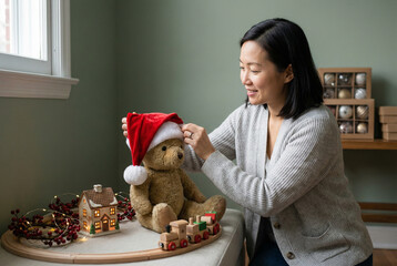 Smiling woman placing festive santa hat on plush bear beside holiday decorations and wooden toy train by bright window