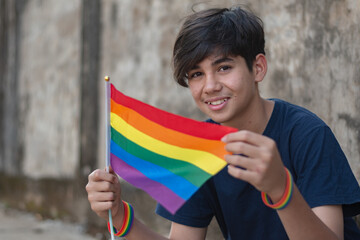 Teenaged boy wears rainbow wristband and holding rainbow flags, lgbtqai symbol, in front of wall, concept for calling out all people to respect human rights of lgbtqai people around the world.