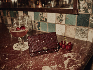 a marble countertop with a silver clutch purse, a cocktail glass with cherries, and a plate of cherries. The background features a mosaic tile backsplash.