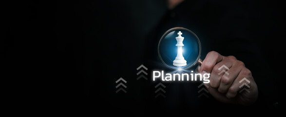 A hand holding a magnifying glass highlighting a chess king with the word planning, representing strategic planning, leadership, decision making, and business growth strategy.