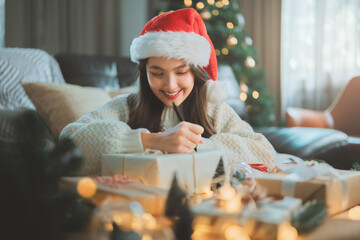 Happy young asian woman in santa hat writing gift tag or wrapping presents in festive living room for christmas and new year holiday preparation