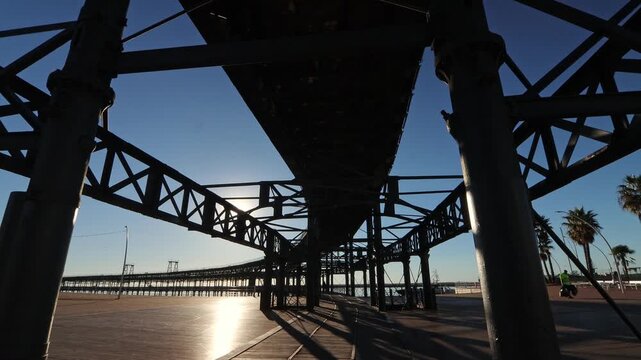Dramatic low angle shot of the historic Muelle del Rio Tinto structure at sunset/sunrise, highlighting its massive ironwork, palm trees, and people enjoying the sunny maritime promenade.