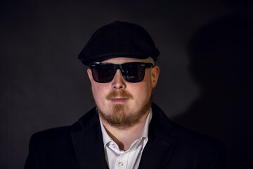 Fototapeta premium Serious adult man wearing sunglasses, flat cap and suit jacket, looking straight at the camera on a dark studio background.