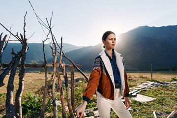 Fototapeta premium Woman in sweater and jacket standing by a rustic fence in countryside mountains at golden sunlight. Outdoor portrait, travel lifestyle with contemplative pose and natural rural scenery.