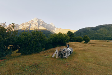 Fototapeta premium Mountain meadow with hiker and backpack sitting on a stonewall, trees and distant peaks at sunrise. Landscape scene of nature, solitude, outdoor travel and hiking adventure.