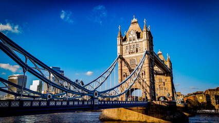 Obraz premium Tower Bridge captured from a low-angle perspective under clear blue skies, highlighting its iconic architecture and the modern London skyline in warm afternoon light.