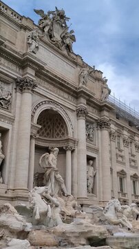 A vertical video pan from top to bottom of the magnificent Trevi Fountain in Rome, Italy, showcasing the Baroque sculptures of Oceanus, the Tritons, and the wild horses, and the turquoise water.